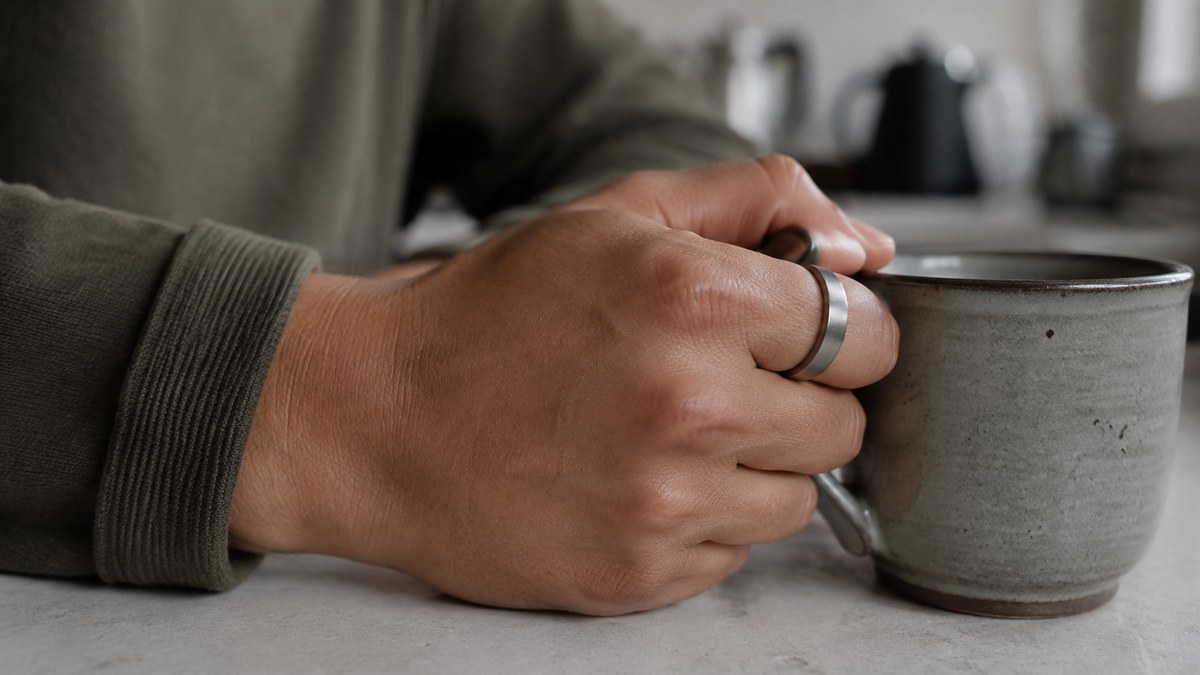 Slim titanium smart ring worn on the index finger while holding a coffee mug